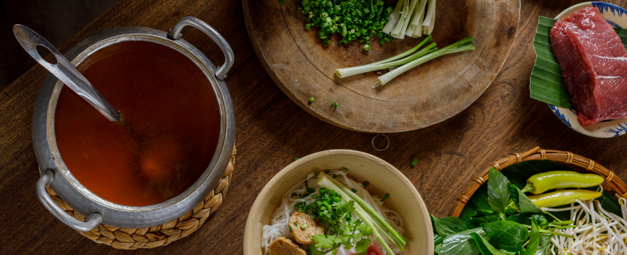 Ein Top mit Pho-Suppe, auf den Tisch sind verschiedene Zutaten.